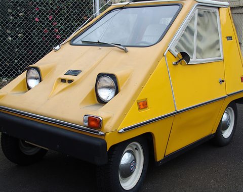 Yellow wedge-shaped electric car with round headlamps in square black surrounds, small indicator lamps and a black bumper, in front of a wire fence. Opens a larger view