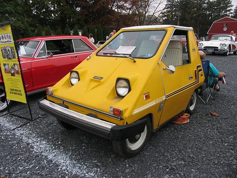 Yellow wedge-shaped electric car with twin round headlamps and a black front bumper on tarmac, a red classic car and an information banner behind. Opens a larger view