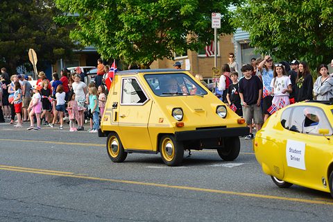 Yellow wedge-shaped electric car driving along a street in a parade, crowds on the pavement and a second small yellow car following. Opens a larger view