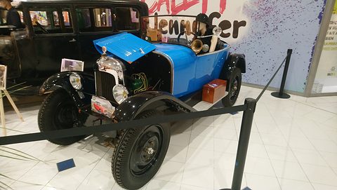 A bright blue tourer with one bonnet side raised and black wings, behind a rope barrier in a mall. Opens a larger view