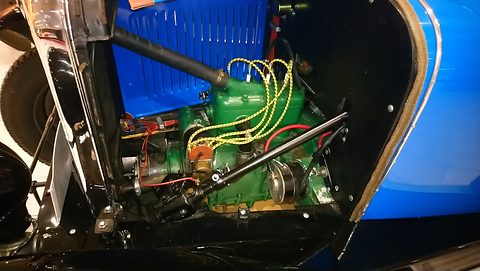 A green-painted four-cylinder engine with yellow plug leads under a raised blue bonnet side. Opens a larger view
