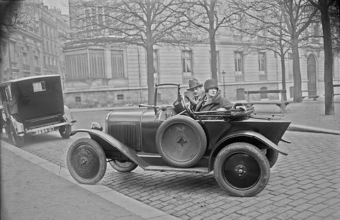 A small open two-seater with a spare wheel on its flank, a man and a woman aboard, on cobbles.