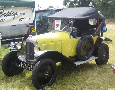 A pale yellow tourer with a black hood and plate PU 5525 on grass before a dealer’s gazebo. Opens a larger view