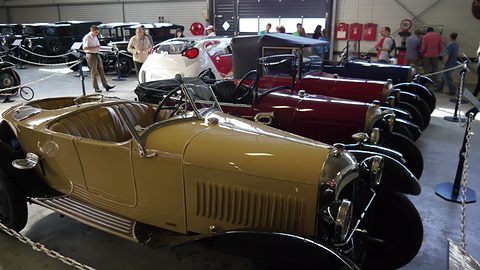A beige open two-seater with a louvred bonnet in a row of red and black Citroëns in a museum hall. Opens a larger view