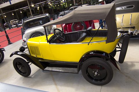 A yellow open two-seater with a grey folding hood and black wings, in a museum hall with lamp posts. Opens a larger view