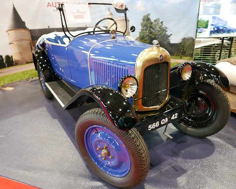 A blue open two-seater with a brass radiator shell and plate 566 QH 42, on a carpeted show stand. Opens a larger view