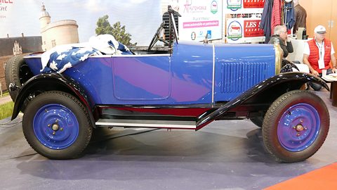 Side view of a bright blue open two-seater with blue wheels and black wings on a show stand. Opens a larger view