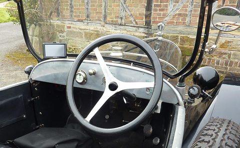 A white-spoked steering wheel before a grey metal dashboard with one round gauge, a brick wall beyond. Opens a larger view