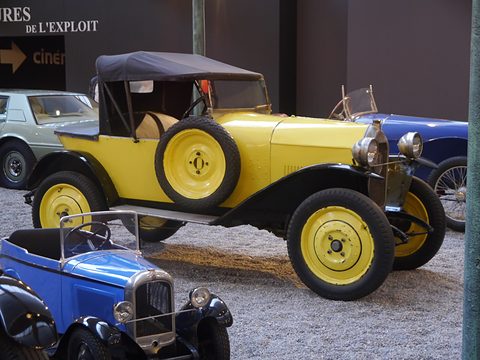 A yellow two-seater with yellow wheels, a spare on its flank and a black hood, a blue small car in front. Opens a larger view