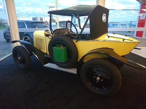 Rear three-quarter view of a yellow two-seater with a pointed tail, a black hood and a spare wheel at the side. Opens a larger view