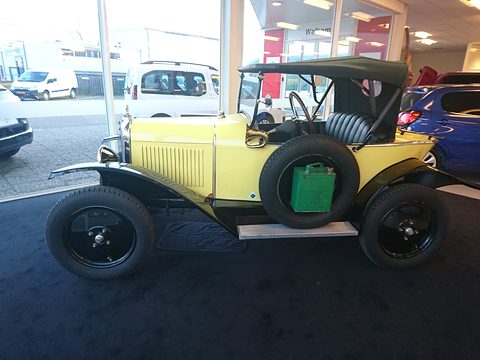 Side view of a pale yellow two-seater with a black hood, a spare wheel and a green petrol can on its flank. Opens a larger view