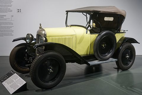 A cream open tourer with a beige hood, black wings and a nickel radiator on a grey museum floor. Opens a larger view