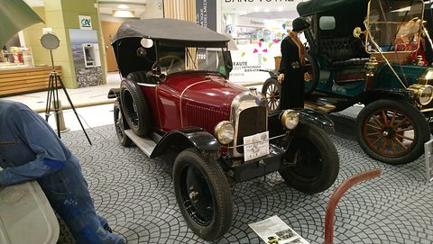 A red tourer with a black hood on a cobble-pattern floor beside a mannequin mechanic and a veteran car. Opens a larger view