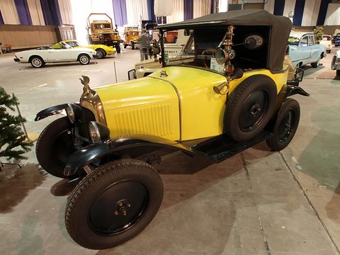A yellow tourer with a black hood and a spare wheel at its side, in a hall with other old cars. Opens a larger view