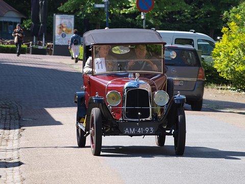 A red car with a black hood and plate AM-41-93 driving toward the camera along a tree-lined street. Opens a larger view