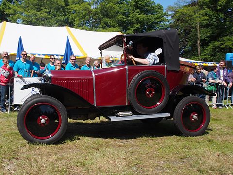 Side view of a red car with a black hood and red wheels moving across grass in front of a crowd. Opens a larger view