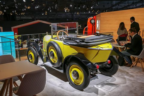 A yellow open car with its hood folded, yellow wheels and a pointed tail beside a stand café. Opens a larger view
