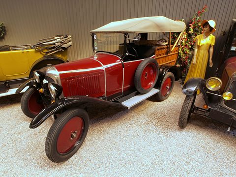 A red tourer with a beige folding hood and red wheels on a gravel floor beside a costumed mannequin. Opens a larger view