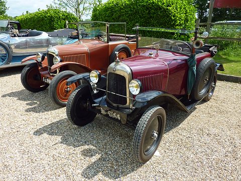 A maroon open car with a black radiator surround parked on gravel beside a brown sister car. Opens a larger view