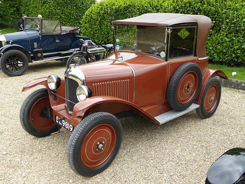 A brown cabriolet with a fabric roof, side screens and brown wheels parked on gravel before a hedge. Opens a larger view