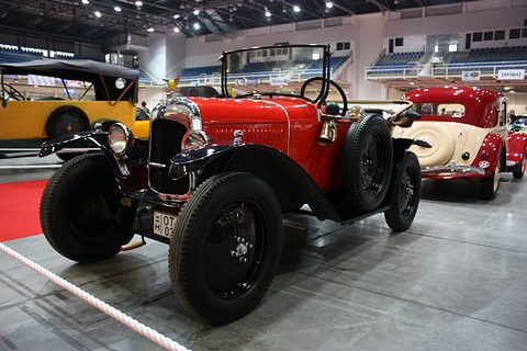 A red open two-seater with black wings and a nickel radiator on a grey floor among other old cars. Opens a larger view