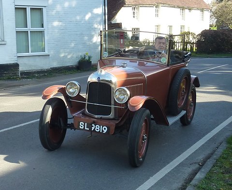 A brown open car with plate SL 9819 being driven along a village road past white houses. Opens a larger view