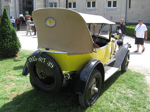 Rear three-quarter view of a yellow car with a beige hood, its tail lid raised over a spare wheel, on grass. Opens a larger view