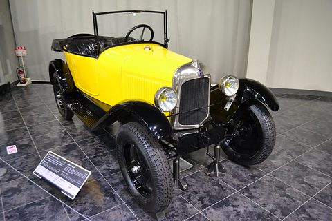 A yellow tourer with black wings and a nickel radiator, its hood folded, beside a museum label. Opens a larger view