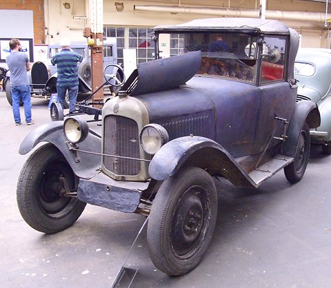 A weathered dark blue car with faded paint, a torn hood and a lifted bonnet side on a concrete floor. Opens a larger view