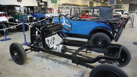 A bare ladder frame on four wheels with an engine, gearbox and steering wheel, a blue car behind. Opens a larger view