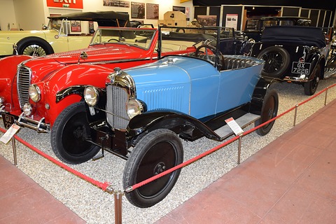 A pale blue open car with black wings behind a red rope, a red sports car beside it. Opens a larger view