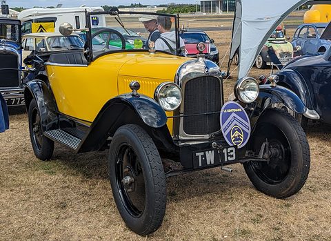 A yellow three-seater with black wings and a Citroën club badge on its bumper, on dry grass. Opens a larger view