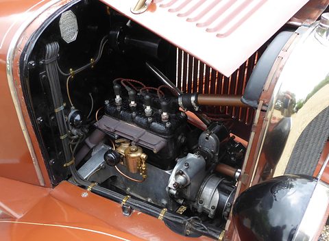 A black four-cylinder side-valve engine with a brass carburettor, plugs and dynamo inside a brown bonnet. Opens a larger view