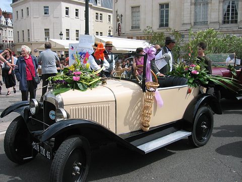 A beige open three-seater decked with flower bouquets, costumed people walking alongside in a town street. Opens a larger view