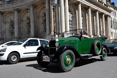 A green open tourer with green wheels parked in a street before a classical stone colonnade, modern cars alongside. Opens a larger view