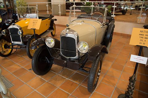 A cream open two-seater with black wings on a terracotta tile floor beside a yellow small car. Opens a larger view