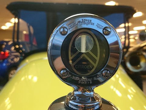 A chrome radiator-cap thermometer with a gauge window above a double chevron badge, the yellow bonnet below. Opens a larger view