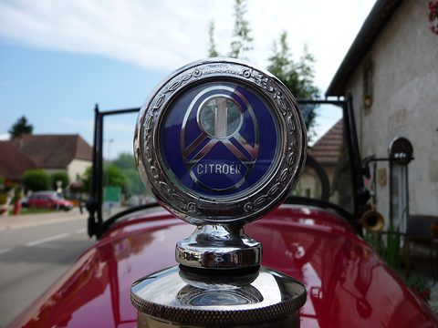 A chrome radiator thermometer with a blue chevron face, a village street blurred behind. Opens a larger view