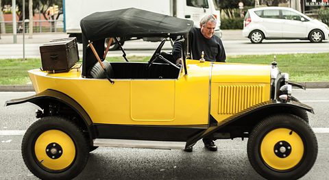 Side view of a yellow car with a black hood and a wooden trunk on its tail, a man leaning on it. Opens a larger view