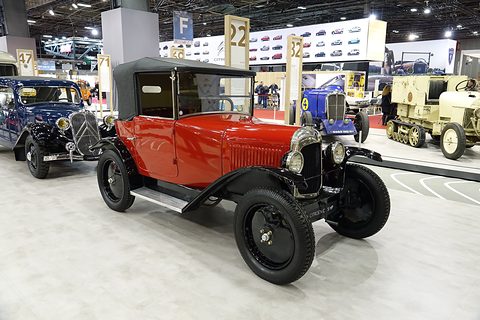 A red cabriolet with a black roof and black wings on a pale show floor, a Traction Avant behind. Opens a larger view
