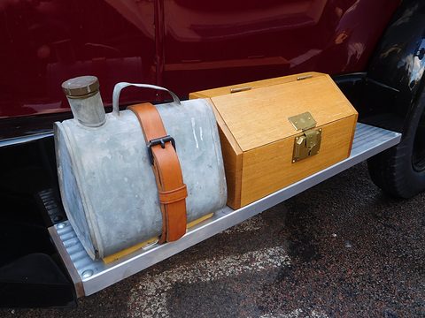 A galvanised petrol can held by a leather strap beside a varnished wooden box on a ribbed running board. Opens a larger view