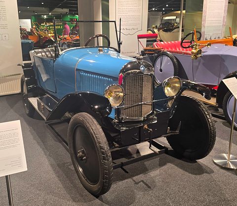 A pale blue open two-seater with black wings, brass-rimmed lamps and a flat radiator, in a museum hall. Opens a larger view