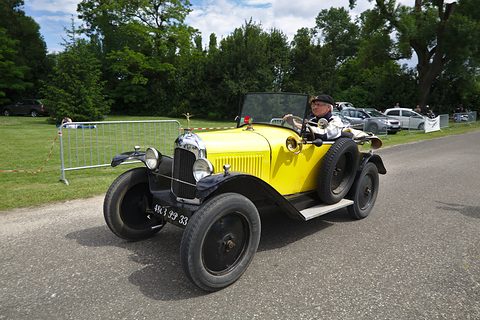 A pale yellow open two-seater with black wings and wheels driving along a park road past parked cars. Opens a larger view
