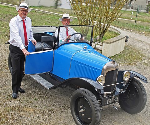 A small blue open tourer with black wings and a chevron radiator, one man at the wheel and another holding the door. Opens a larger view