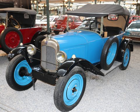 A light blue open three-seater with a black hood and light blue wheels on a gravel museum floor. Opens a larger view