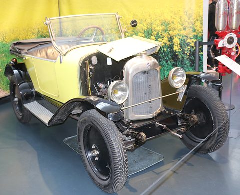 A pale yellow open car with a nickel radiator and one bonnet side raised, before a painted field backdrop. Opens a larger view