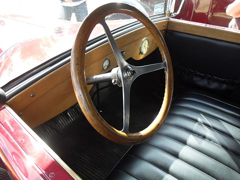 A wood-rimmed steering wheel before a varnished wooden dashboard and pleated black leather seats. Opens a larger view