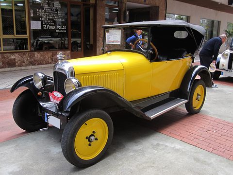 A yellow tourer with yellow wheels, black wings and a black hood on a red-paved street before shopfronts. Opens a larger view