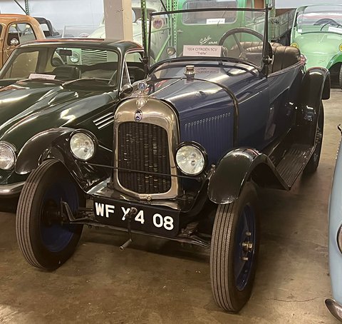 A dark blue open three-seater with a nickel radiator and a starting handle, among other cars in a museum. Opens a larger view
