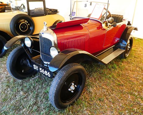 A red open three-seater with black wings, brass horn and plate SY 5686 on dry grass inside a marquee. Opens a larger view